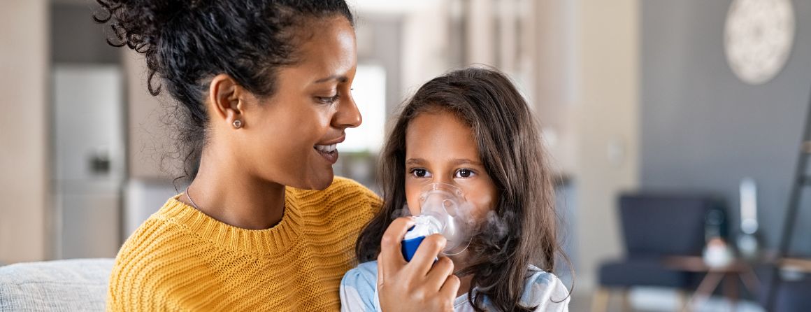 Mother with yellow knit jumper helping daughter with inhaling through a nebulizer. 