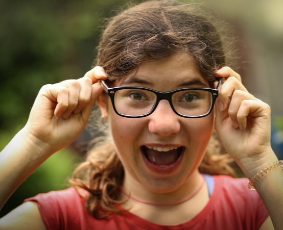 Little girl smiles at the camera while holding her black rimmed glasses